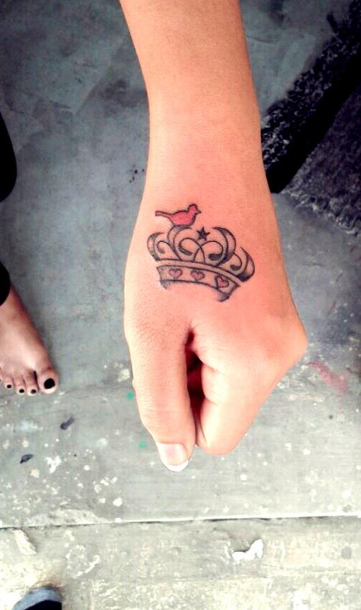 A feminine crown with pink bird embellishments in this tattoo on hand.