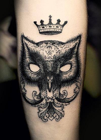 Black and grey face of an owl is embellished with a hovering crown on forearm.