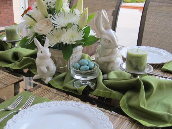 Natural spring tablescape decorated with linens, plates, glassware, flowers, candles and decorative items such as ceramic white bunnies and moss eggs.