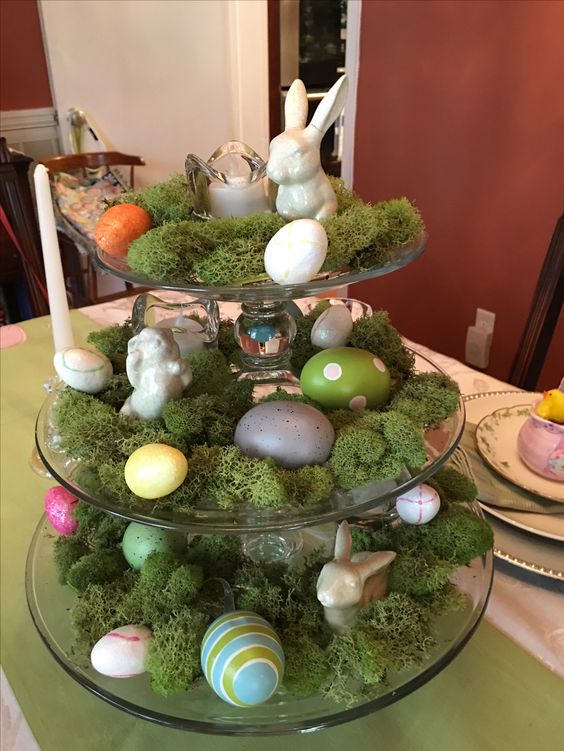 Tiered tray centerpiece decorate with fake moss, plastic eggs and a porcelain bunnies.